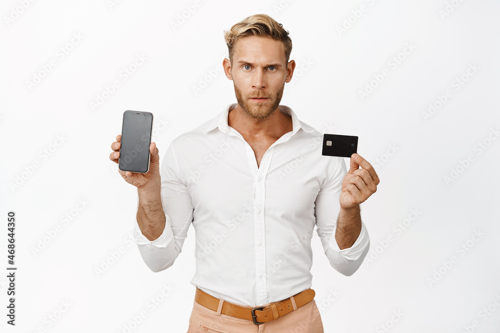 Serious blond man showing credit card with smartphone app screen, standing over white background Serious blond man showing credit card with smartphone app screen, standing over white background