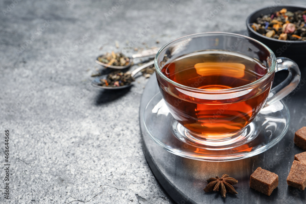 Glass cup of freshly brewed tea, dry leaves and sugar on grey table. Space for text Glass cup of freshly brewed tea, dry leaves and sugar on grey table. Space for text