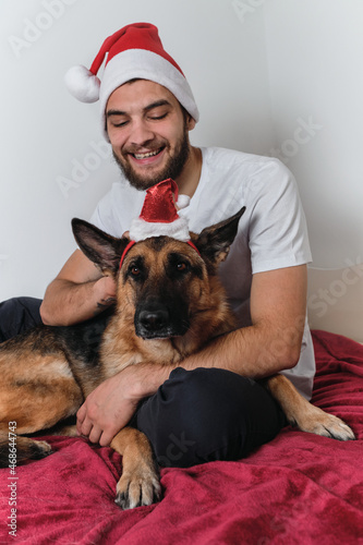 Celebrate Christmas and New Year together with dog at home. Young Caucasian European man with beard and red Santa hat is sitting on blanket and hugging German Shepherd.