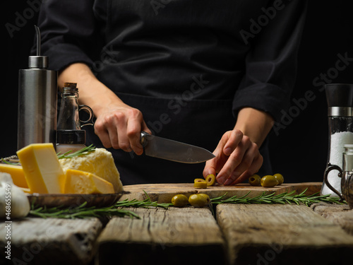 The chef is cutting green olives on a cutting board. Ingredients for making salad, pizza, focaccia. Wooden texture. Restaurant, hotel, cafe, cookbook, home cooking.