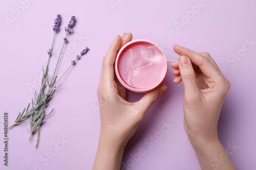 Canvastavla Woman taking under eye patch with spatula out of jar near lavender flowers on lilac background, top view