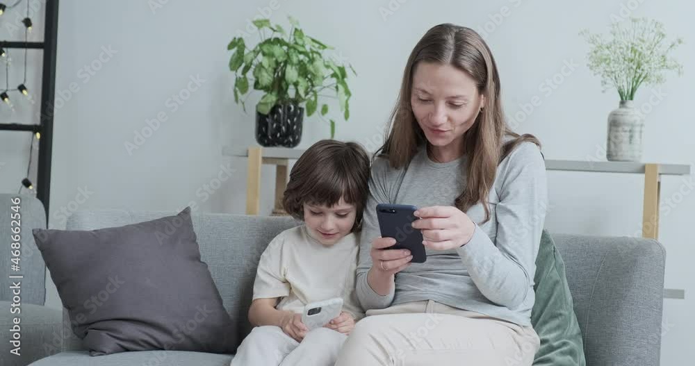 Laughing woman texting typing message on telephone having positive emotion. Smiling male kid playing online internet game use smartphone. Modern family relaxing on couch with digital electronic device Laughing woman texting typing message on telephone having positive emotion. Smiling male kid playing online internet game use smartphone. Modern family relaxing on couch with digital electronic device