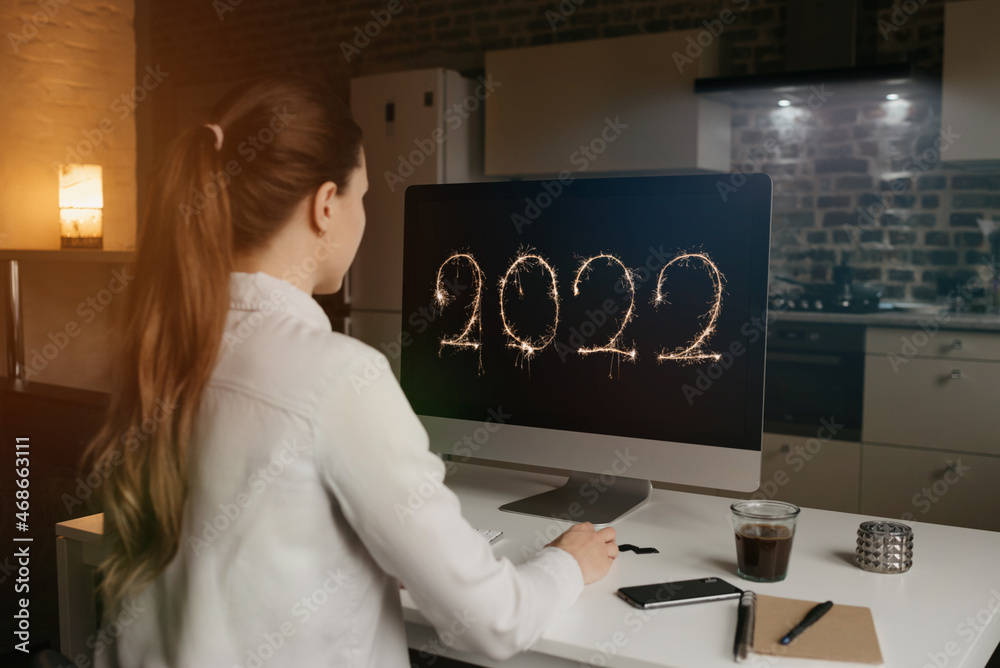 A back view of a woman who is staring at the number 2022 on a display of her desktop computer at home. A businesswoman is watching a new year video. - obrazy, fototapety, plakaty 