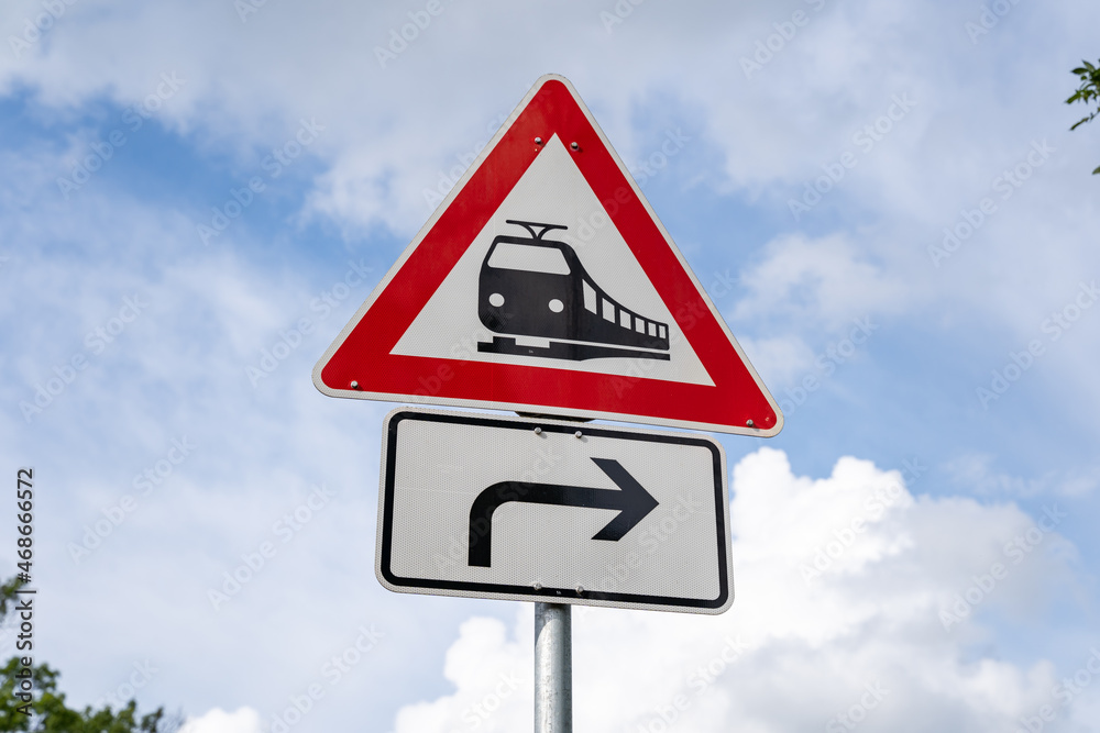 Rail crossing without barriers ahead traffic sign in Germany. Warning symbol to pay attention in front of the sky. A black train on a white triangle with a red outline. An arrow pointing to the right. Rail crossing without barriers ahead traffic sign in Germany. Warning symbol to pay attention in front of the sky. A black train on a white triangle with a red outline. An arrow pointing to the right.