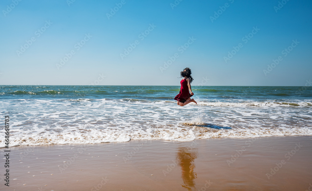Young woman of indian ethnicity joyfully jumping on paradise beach of Goa ( Unrecognizable person ) - obrazy, fototapety, plakaty 