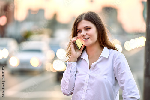 Telephone conversation near highway with heavy traffic, young woman uses smartphone on city street on summer evening.