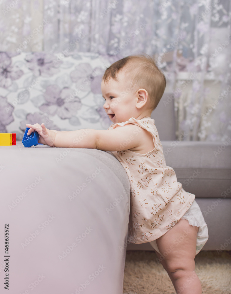 Fototapeta premium Adorable baby stands near the sofa and plays with colored toys