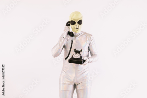 Person dressed in silver suit and green alien mask, talking on a black retro telephone from the 60s, on white background
