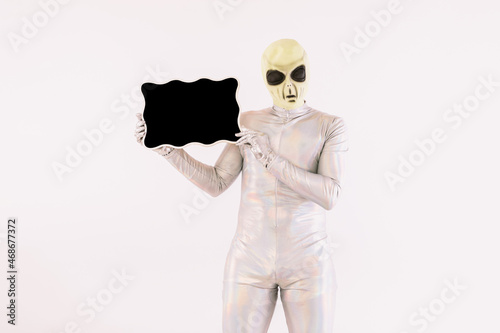 Person dressed in silver suit and green alien mask, holding a blackboard, on white background