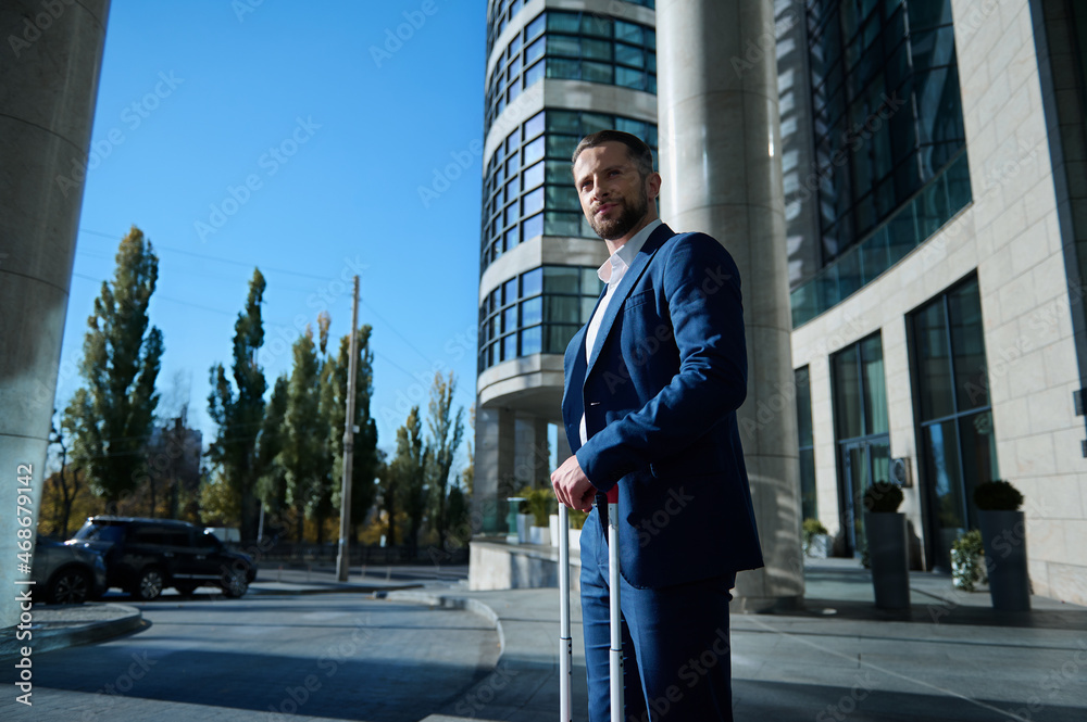 Bottom view of a self-confident successful businessman developer entrepreneur in business suit and luggage smiles looking aside standing outdoor on modern high-rise building glass entrance background Bottom view of a self-confident successful businessman developer entrepreneur in business suit and luggage smiles looking aside standing outdoor on modern high-rise building glass entrance background