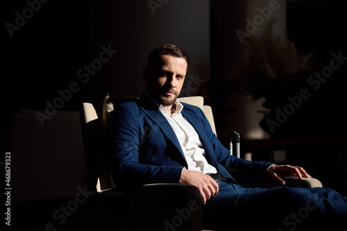 Attractive self-confident business traveler, handsome successful businessman, entrepreneur resting in armchair in hotel lobby and confidently looking at camera