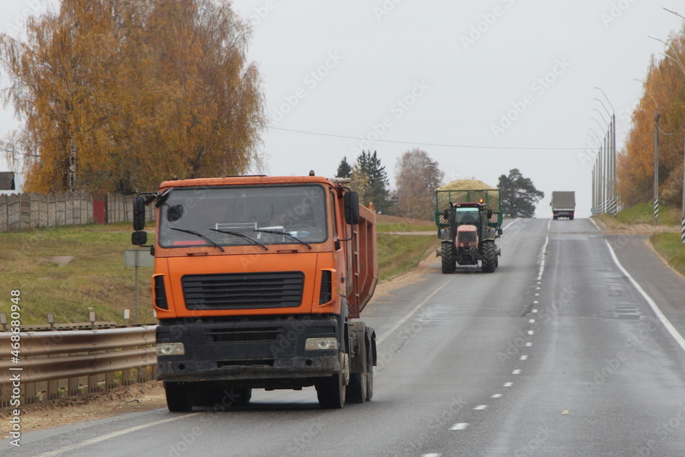 Yellow dump truck drive on asphalted road on tractor and yellow trees background at autumn day, Russian farming logistics