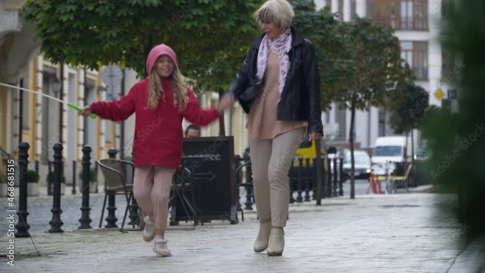 Wide shot happy cheerful girl with balloon walking jumping holding hands with granny outdoors. Excited Caucasian granddaughter enjoying weekend with grandmother on city street having fun Wide shot happy cheerful girl with balloon walking jumping holding hands with granny outdoors. Excited Caucasian granddaughter enjoying weekend with grandmother on city street having fun