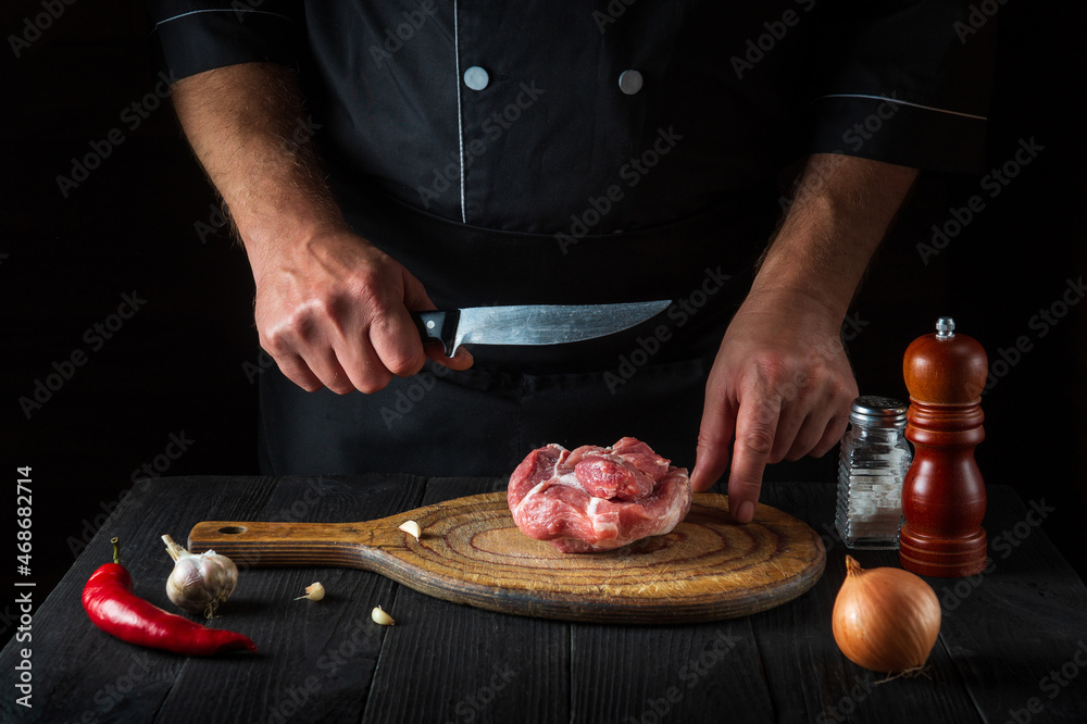 Professional chef cuts meat with a knife in the kitchen prepares food. Vegetables and spices on kitchen table in a restaurant to prepare delicious lunch.