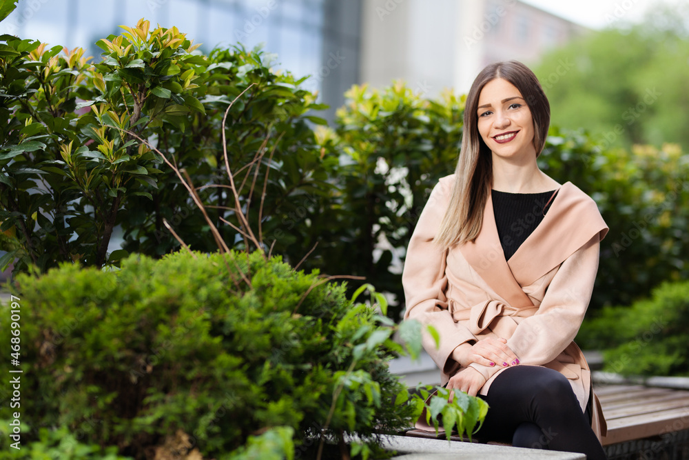 Portrait of a woman sitting on a bench. Successful businesswoman posing outdoors surrounded by green plants. Portrait of a woman sitting on a bench. Successful businesswoman posing outdoors surrounded by green plants.