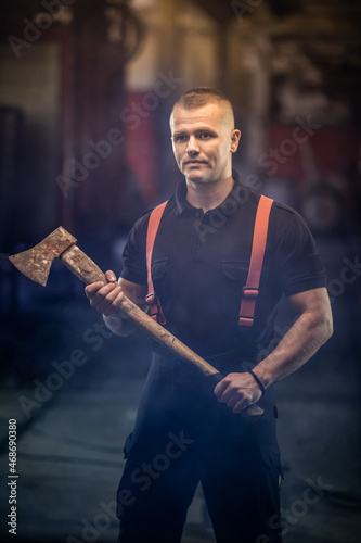 firefighter portrait wearing shirt and red throuser suspenders, holding an axe. smoke and fire trucks in the background.
