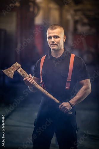 firefighter portrait wearing shirt and red throuser suspenders, holding an axe. smoke and fire trucks in the background. photo