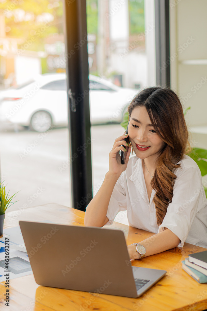 Smiling Asian businesswoman in the modern office using laptop and graph paper to analyze financial calculations while sitting at her desk in modern office. Smiling Asian businesswoman in the modern office using laptop and graph paper to analyze financial calculations while sitting at her desk in modern office.