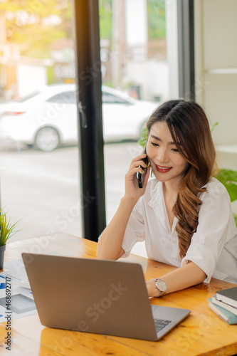 Smiling Asian businesswoman in the modern office using laptop and graph paper to analyze financial calculations while sitting at her desk in modern office.