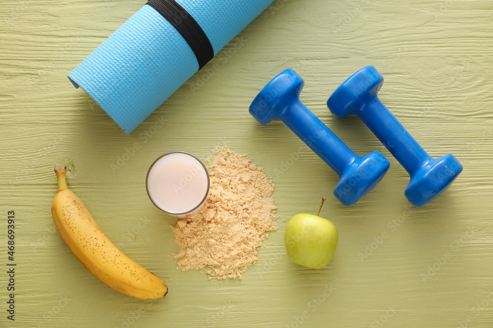 Glass of protein shake, powder, banana, apple, dumbbells and fitness mat on color wooden background Glass of protein shake, powder, banana, apple, dumbbells and fitness mat on color wooden background