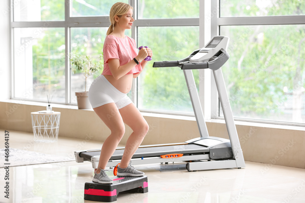 Young pregnant woman training with aerobic stepper and dumbbells in gym