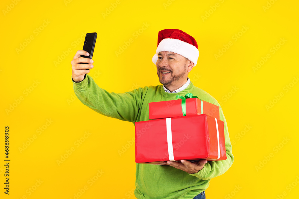 Portrait of senior latin man holding mobile phone taking a photo selfie with copy space in a christmas concept on yellow background