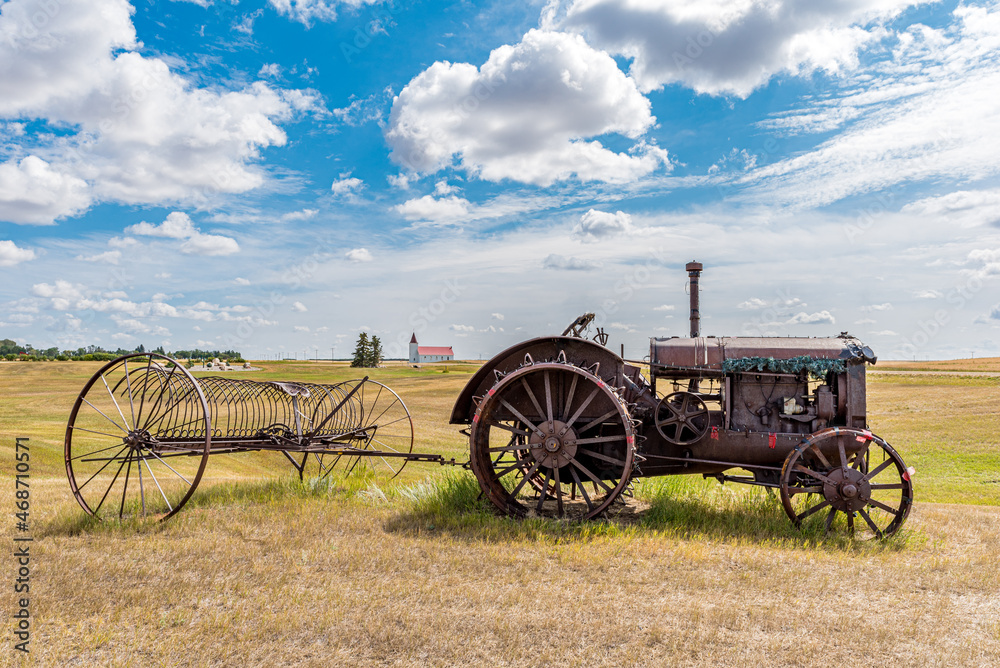 Antique tractor with steel wheels and lugs pulling a hay rake with St. Anthony Roman Catholic Church in Illerbrun, SK in the background