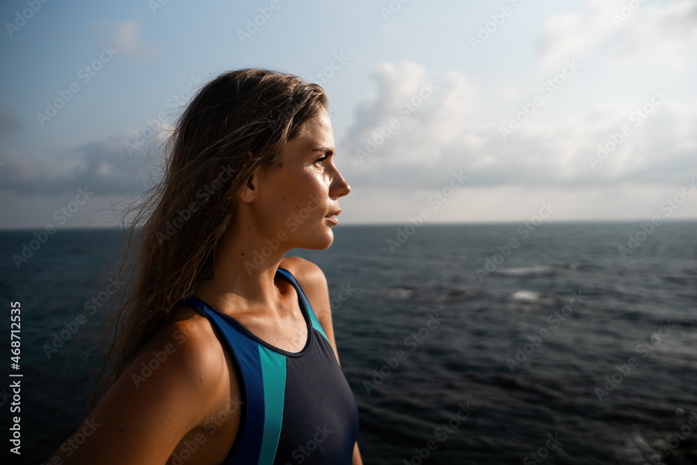 A young beautiful tanned woman with long hair in blue bikini stands at rock near blue green clear ocean sea water. Woman relax on sunset at sea, enjoy holidays and weekend vacation in summer time.