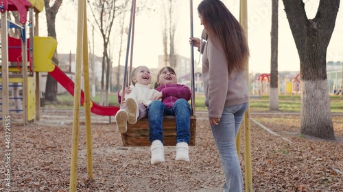 Happy young mother and little daughters swing on a swing in the park in autumn. Beautiful little girls sit on a swing and laugh. Mom, the children play together in the playground. Happy family concept