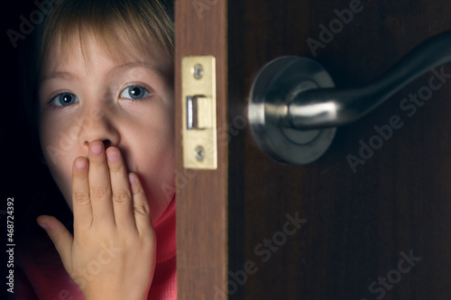 Photos A little girl, a child, peeks out of the ajar door and covers her mouth with her hand in surprise and fear