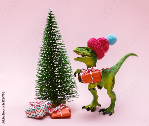Fototapeta Naklejka Na Ścianę i Meble -  Funny plastic toy dinosaur wearing knitted hat with  present boxes and evergreen tree on a pink background.