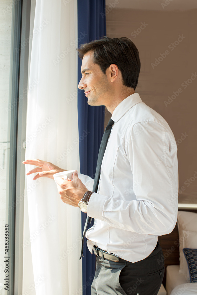 Handsome young businessman drinks coffee in front of the hotel window Handsome young businessman drinks coffee in front of the hotel window