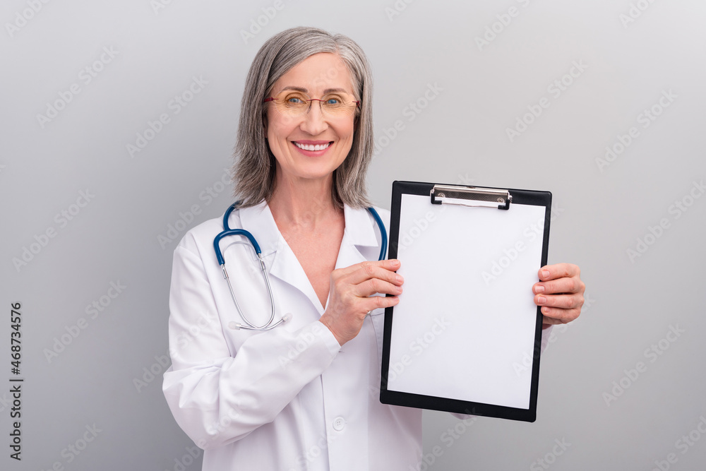 Photo of positive good mood senior woman doc dressed white uniform glasses holding clipboard empty space isolated grey color background Photo of positive good mood senior woman doc dressed white uniform glasses holding clipboard empty space isolated grey color background