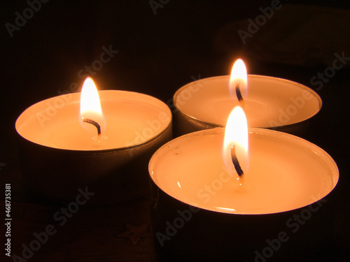 Three burning tealight s with wick and flame against a black background. Close-up, focus on foreground.