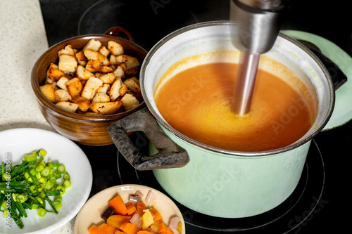 Hand blender in pot mixes soup, vegetable and fried piece of bread on board. photo