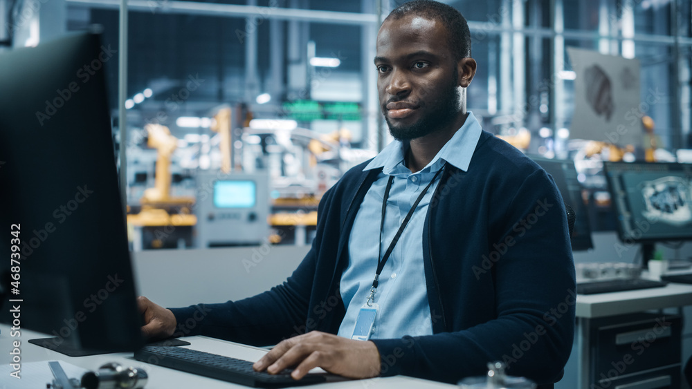 Car Factory Office: Portrait of Confident Black Male Chief Engineer Working on Desktop Computer. Professional Technician in Automated Robot Arm Assembly Line Manufacturing High-Tech Electric Vehicles