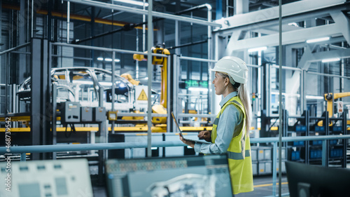 Car Factory: Female Automotive Engineer Wearing Hard Hat, Standing, Using Laptop. Monitoring, Control, Equipment Production. Automated Robot Arm Assembly Line Manufacturing Electric Vehicles. © Gorodenkoff