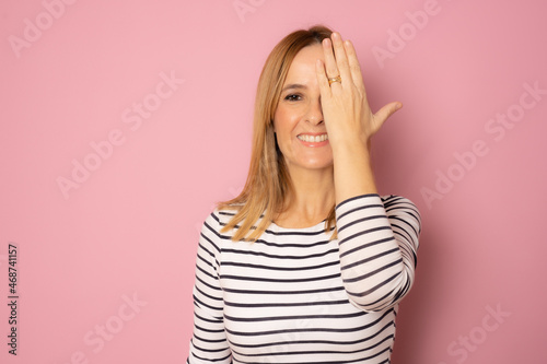 Young beautiful blonde woman wearing casual t-shirt standing over pink isolated background covering one eye with hand, confident smile on face and surprise emotion.