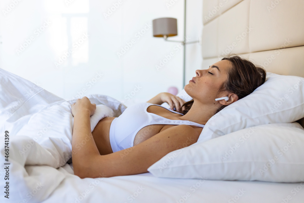 Young beautiful woman relaxing in her bed, she is listening to music and lying down with eyes closed. Woman Lying in Bed and Listening to Music Through her Wireless Earphones Young beautiful woman relaxing in her bed, she is listening to music and lying down with eyes closed. Woman Lying in Bed and Listening to Music Through her Wireless Earphones