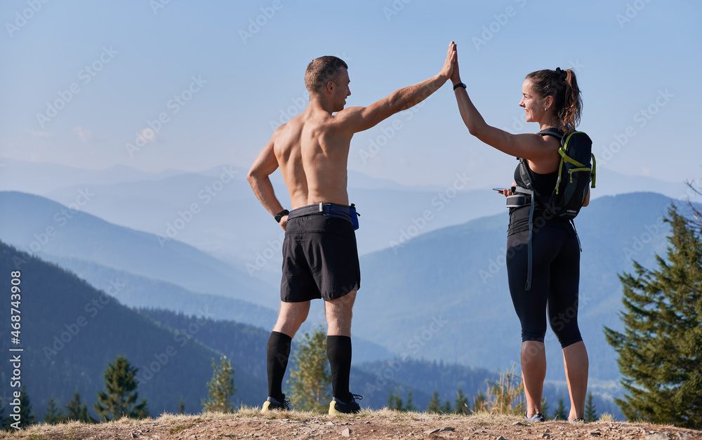 Rear view of two trail runners clapping their hands while standing on top of hill on sunny day, against the backdrop of incredible mountain scenery. Rear view of two trail runners clapping their hands while standing on top of hill on sunny day, against the backdrop of incredible mountain scenery.