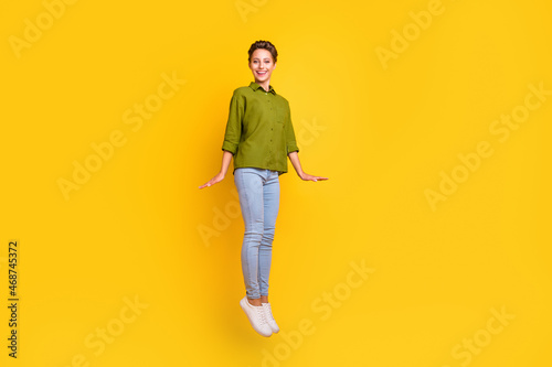 Photo of carefree cheerful lady jump shy pose wear green shirt jeans sneakers isolated yellow color background