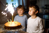 Two beautiful kids, little preschool boys celebrating birthday and blowing candles on homemade baked cake, indoor. Birthday party for siblings children. Happy twins about gifts and fireworks on tarte.
