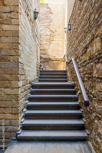 Architectural detail of outdoor wooden old weathered narrow staircase going up with wooden handrail  between two stone bricks walls  in abandoned building