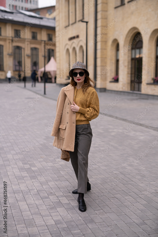 Fototapeta premium Charming model girl in an autumn beige coat and sunglasses, plaid panama hat standing with face turned holding coat on her shoulders. Retro toned photo
