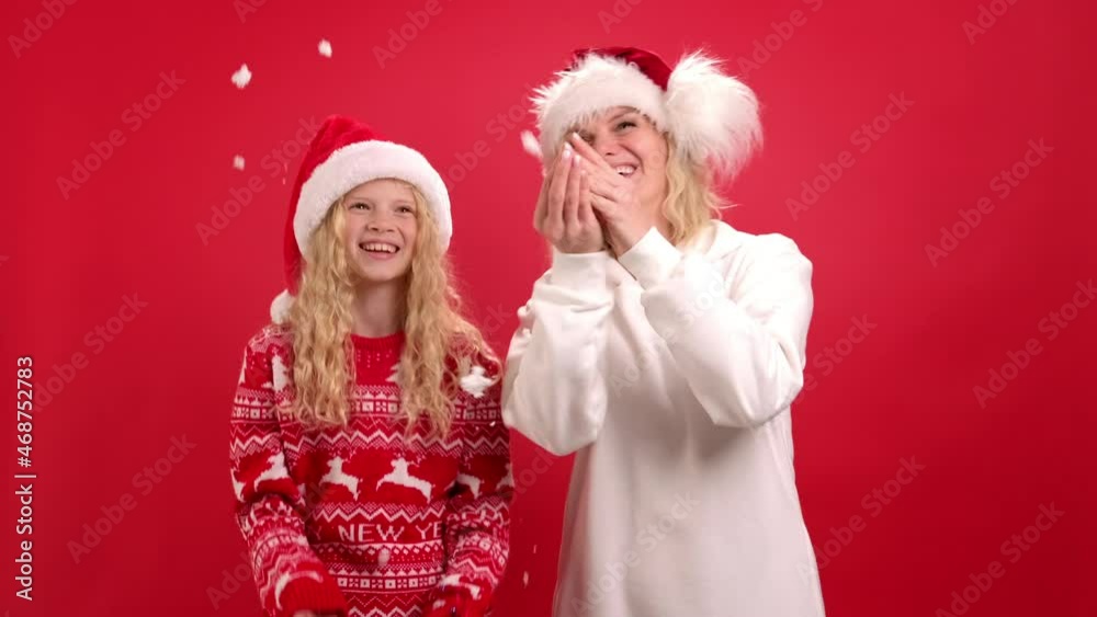 Christmas Holidays Portrait happy family of two In Santa hats mother and daughter having fun, throwing snow and blowing snow off their hands on red studio background. Christmas Holidays Portrait happy family of two In Santa hats mother and daughter having fun, throwing snow and blowing snow off their hands on red studio background.