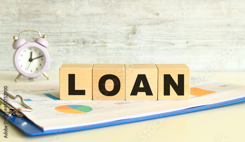 Wooden cubes lie on a folder with financial charts on a gray background. The cubes make up the word LOAN.