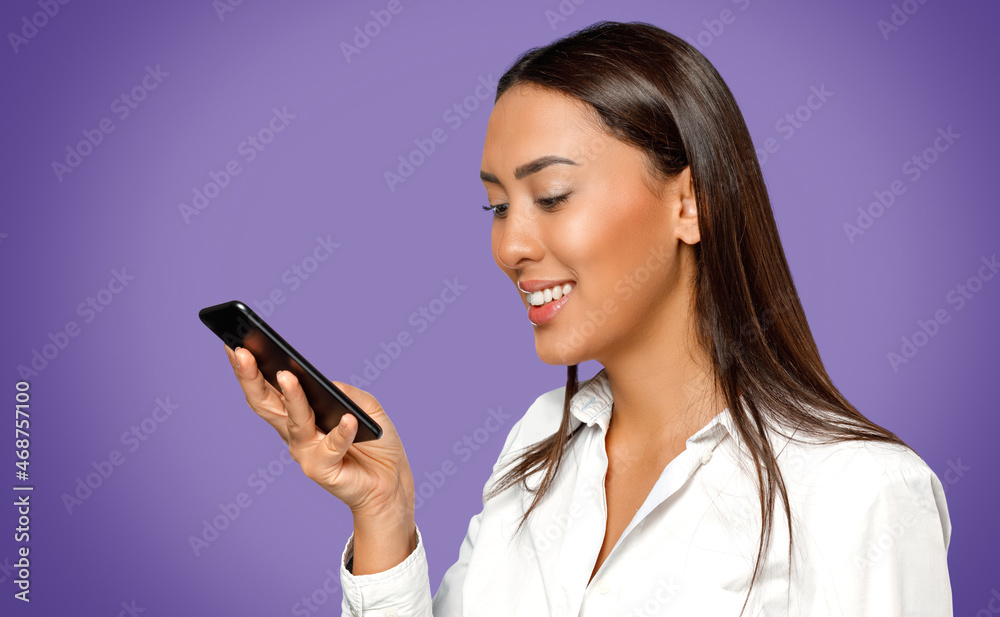 Headshot portrait of smiling young woman wearing white shirt and looking at her mobile phone isolated on purple coloured background. Headshot portrait of smiling young woman wearing white shirt and looking at her mobile phone isolated on purple coloured background.