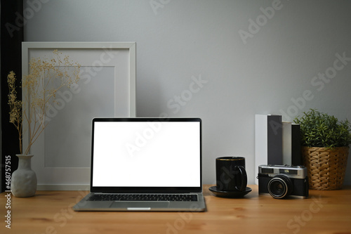 Photo of a white blank screen computer laptop on the wooden table surrounded by a coffee cup  vintage camera  empty picture frame potted plant and books.