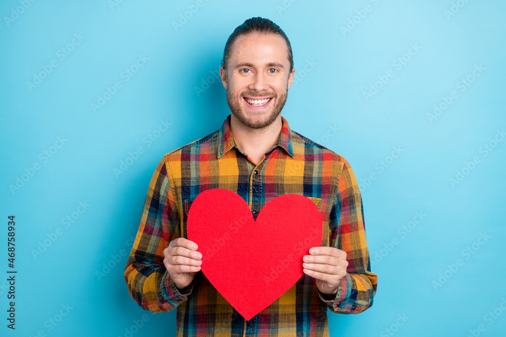 Photo of cute millennial tail hairstyle guy hold heart wear plaid shirt isolated on blue color background Photo of cute millennial tail hairstyle guy hold heart wear plaid shirt isolated on blue color background