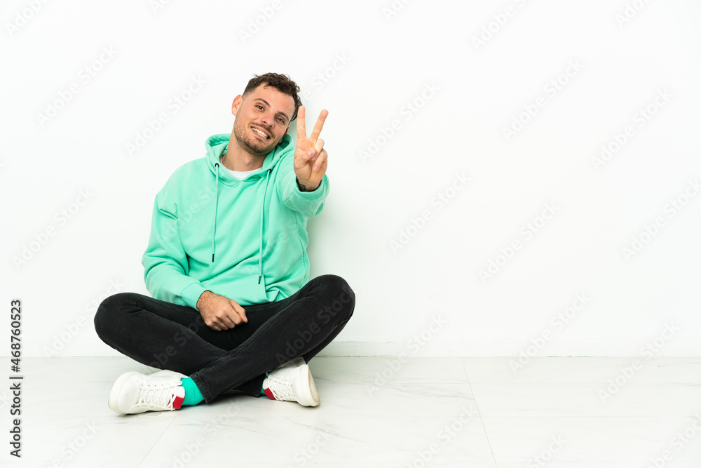 Young handsome caucasian man sitting on the floor smiling and showing victory sign Young handsome caucasian man sitting on the floor smiling and showing victory sign